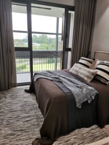 Hotel bedroom with a large glass balcony door, brown bedspread, gray throw, and striped pillows on the bed.