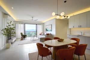 Open-plan living/dining area with a round marble dining table and orange chairs, gray sofa, potted plant, and a large window view outside.
