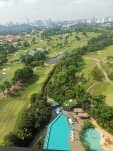 Aerial view of a lush golf course with ponds, winding fairways, and dense trees, plus a resort pool area in the foreground and a distant city skyline.