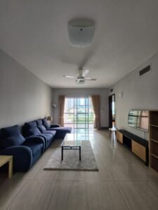 Living room with a dark blue sectional sofa along the left wall, a glass coffee table on a light rug, and a TV stand on the right; a glass door leads to a balcony with city buildings outside.