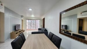 Open-plan living and dining area with a wooden dining table, black upholstered chairs, and a TV on a low stand facing a gray sofa; balcony doors at the far end.