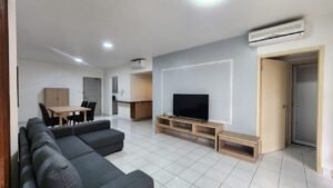 Spacious living room with a gray sectional sofa, a TV on a wooden console, and a dining table with four chairs; open kitchen in the background pale floor tiles.