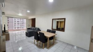 Open-plan living and dining area with a gray L-shaped sofa, wooden dining table and six black chairs, near sliding glass balcony doors.