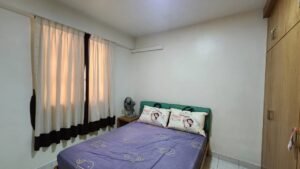 Small bedroom with a double bed, purple elephant-pattern sheets, two pillows, a table fan, cream curtains, and a wooden wardrobe.