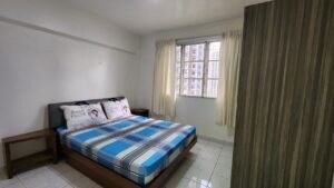 Bedroom with a double bed, blue plaid sheets, and two decorative pillows near a small wooden nightstand under a window with cream curtains
