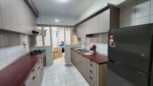Narrow kitchen with beige cabinets, dark red countertops, gas stove left, double sink right, fridge at far end; laundry area visible through doorway.