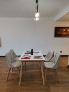 Modern dining room with a white rectangular table set for six, surrounded by light gray upholstered chairs, red napkins and white dishes, a purple centerpiece, wooden floor, and a pendant light above.