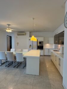 Bright white kitchen with a central island, glass dining table, and white chairs under yellow pendant lights.
