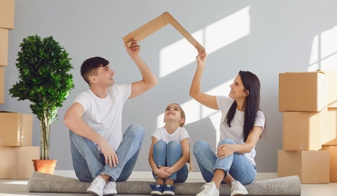 A family of three sits on a rug among moving boxes, smiling as they hold up a cardboard piece together.