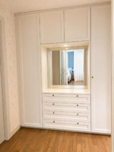 White built-in wardrobe with a central mirrored panel, four drawers below, and tall cabinets on the sides in a bedroom setting as hardwood flooring shows below.