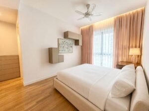 Bright modern bedroom with a low beige bed, white bedding, wooden floor, wall-mounted shelves, a framed art piece, and a window with sheer and tan curtains next to a nightstand and lamp.