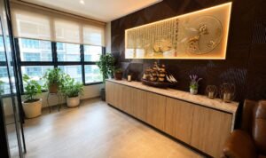 Bright office corner with a long wooden cabinet, illuminated wall art, a model ship, candles, and green plants by the windows.