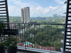 Balcony railing with potted plants overlooking a cityscape of tall buildings and dense green trees, with a blue pool visible below.