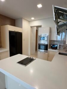 Modern white kitchen with island cooktop, built-in oven and microwave, and a tall black refrigerator.
