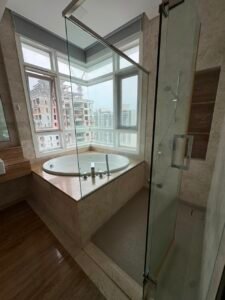 Corner bathroom with a built-in oval bathtub by large windows overlooking city high-rises; glass shower enclosure to the right.