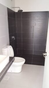 Bathroom with dark tiled shower area, rainfall showerhead, white toilet, and sink countertop on gray floor tiles.