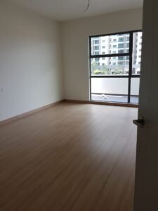 Empty unfurnished room with light wood flooring and a large window/view of a neighboring high-rise building. Ceiling light wiring hanging down.