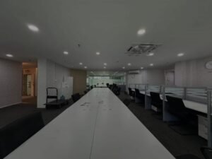 Long white conference table in an empty open-plan office with rows of black chairs and glass partitions on the right, ceiling lights above.