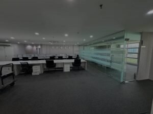 Empty modern office with long white desks and black chairs in an open-plan space, glass partitions on the right.