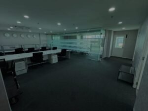 Open-plan office with long white desks, black chairs, and a glass-walled meeting area in the background