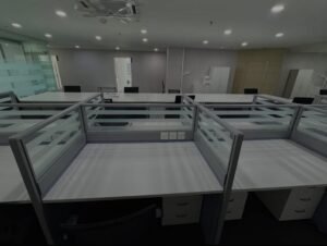 Open-plan office with gray desks and glass partition cubicles, empty workspace under bright ceiling lights.