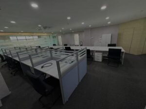 Open-plan office with several white desks and black chairs, glass partition cubicles, and bright ceiling lights.