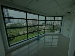 Corner office or balcony-like room with expansive curved windows, showing a city skyline and greenery outside; a large blue water jug sits on the glossy tiled floor inside.
