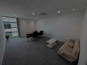 Bright minimalist office with a long desk, two black chairs, and a beige sofa near a large window with a potted plant nearby.
