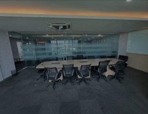 Modern conference room with a large oval table and multiple black office chairs, glass partition walls, and a city view outside.