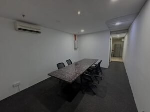 Meeting room with a long marble table and several black chairs, white walls, gray carpet, and a wall-mounted AC; hallway leads to a kitchenette with a fridge in the back.