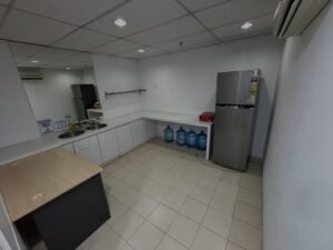 Small white kitchenette with a double sink, white cabinets, and a stainless fridge; several large water jugs under the counter and a wooden island in the foreground.