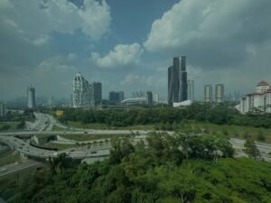 City skyline with modern high-rise buildings and a multi-level highway cutting through green hills under a cloudy sky.