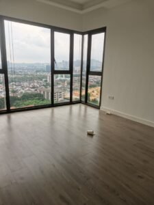 Empty, unfurnished room with a corner of floor-to-ceiling windows showing a distant cityscape and construction site; a couple of paper cups lie on the light wood floor.