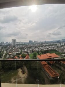 Balcony view of a cityscape with a glass railing, red-tiled roofs, low houses, and distant high-rise buildings under a cloudy sky.