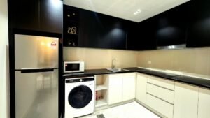 Modern L-shaped kitchen with black glossy upper cabinets, white lower cabinets, beige backsplash, and stainless steel refrigerator, microwave, and washing machine beneath the counter.