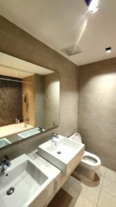 Modern bathroom with beige wall tiles, a double rectangular sink vanity, and a large mirror; toilet on the right and a bathtub with a mosaic-tile wall in the back.