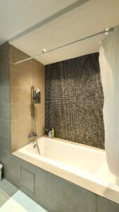 White bathtub with a handheld shower in a tile-surrounded bathroom; a black mosaic wall contrasts with beige side walls and a partially drawn white shower curtain.